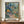 May include: Framed art print in warm wood frame displayed on wooden sideboard alongside white ceramic vase and stacked books. The colorful botanical artwork features vibrant tropical foliage, palm trees, flowers in coral and yellow tones, and a small figure on a winding path through lush vegetation. The piece has layered, painterly textures with blues, greens, oranges and purples creating a dreamy garden scene against cream gallery wall with white molding.