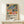 May include: Framed poster with colorful Matisse-style botanical artwork featuring abstract plants, fruits, and leaves in blues, oranges, pinks, and greens, displayed on light wood shelf next to small wrapped gift box with natural twine and greenery sprig against cream wall