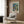 May include: Framed botanical art print with tropical garden scene in blues, greens, and coral colors, displayed on wood-paneled wall above beige upholstered armchair with geometric patterned pillow. Golden floor lamp and dark side table with open book visible in modern neutral-toned room setting.