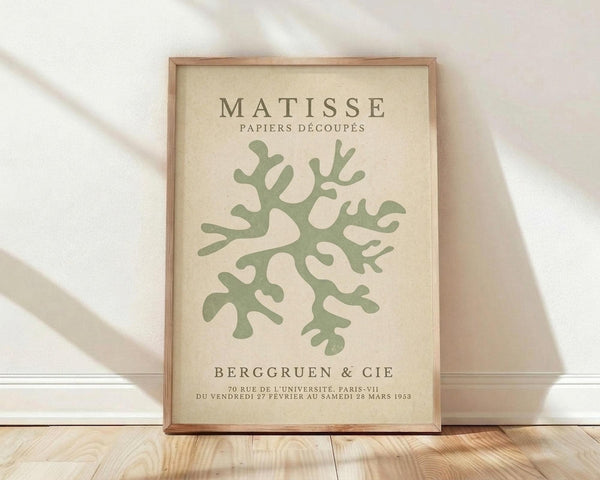 May include: Framed art print leaning against white wall on light wooden floor. Cream-colored poster features green coral-like botanical cutout design in Matisse paper cut style. Text reads "MATISSE PAPIERS DÉCOUPÉS" at top and "BERGGRUEN & CIE" with Paris gallery details below. Natural wood frame with warm lighting and soft fabric shadows visible in background.