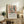 May include: Framed Matisse-style poster with colorful still life artwork showing flowers in vase, fruit bowl, and window scene in muted orange, green, and coral tones. Displayed on rustic wood sideboard with fern plant, ceramic vases, stacked books, and cream upholstered chair in cozy living room setting with white walls and warm natural lighting.