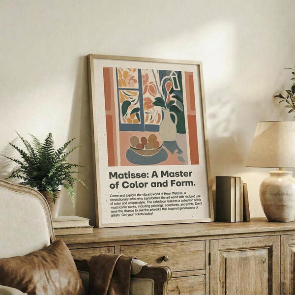 May include: Framed Matisse-style poster with colorful still life artwork showing flowers in vase, fruit bowl, and window scene in muted orange, green, and coral tones. Displayed on rustic wood sideboard with fern plant, ceramic vases, stacked books, and cream upholstered chair in cozy living room setting with white walls and warm natural lighting.