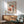 May include: Framed Matisse exhibition poster with colorful abstract floral cutout designs in coral, pink, and black tones, displayed above mid-century modern wooden credenza with white ceramic lamp, next to sage green upholstered armchair with wooden frame in sunlit room with white walls