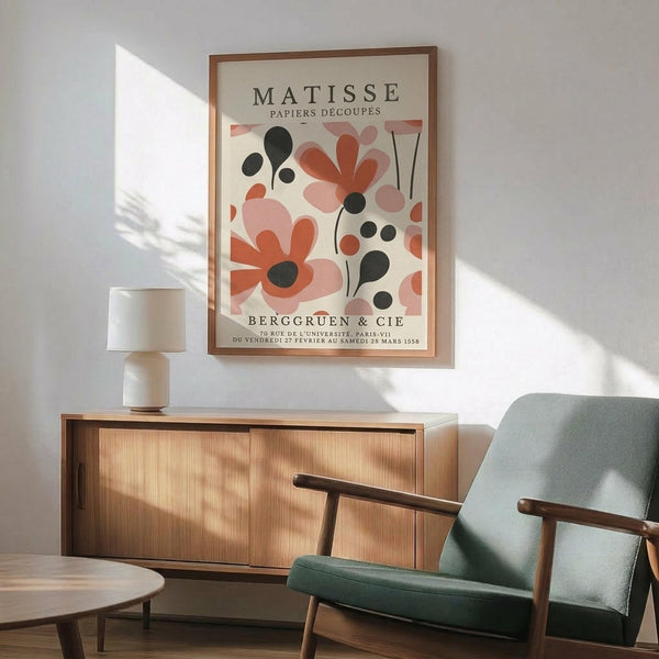 May include: Framed Matisse exhibition poster with colorful abstract floral cutout designs in coral, pink, and black tones, displayed above mid-century modern wooden credenza with white ceramic lamp, next to sage green upholstered armchair with wooden frame in sunlit room with white walls