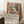 May include: Framed art poster leaning against white wall on wooden floor next to coffee cup and saucer. Artwork shows colorful interior scene with window, vase of flowers, and fruit bowl in warm earth tones of orange, green, and cream. Exhibition-style poster includes descriptive text below the artwork. Natural lighting creates soft shadows across the cozy room setting with textured rug visible.