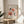 May include: Framed vintage-style Matisse exhibition poster featuring abstract floral cutout designs in coral pink, orange, and dark green shapes on cream background. Displayed on light wood round side table next to beige upholstered chair with plant leaves visible, soft natural window lighting creating shadows on white wall.