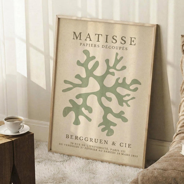 May include: Framed vintage-style art print featuring green organic coral-like shape on cream background, displayed in light wood frame leaning against white wall. Coffee cup on wooden side table and textured white rug visible. Natural lighting creates soft shadows in minimalist room setting.