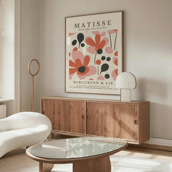 May include: Cream-colored poster with coral pink and black abstract floral shapes hanging above a wooden credenza in a minimalist living room. White curved chair and glass coffee table visible in foreground. Poster features botanical cutout-style designs with French text at top and bottom.