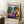 May include: Framed Matisse exhibition poster featuring a woman in purple patterned dress with orange headwrap, reclining among tropical plants in terracotta pots. Vibrant blues, greens, oranges in fauve style. Cream background with "MATISSE" in bold purple letters, "06200 NICE, FRANCE" at bottom. Light wood frame leaning against white wall next to beige sofa, with coffee cup on wooden side table and white textured rug.