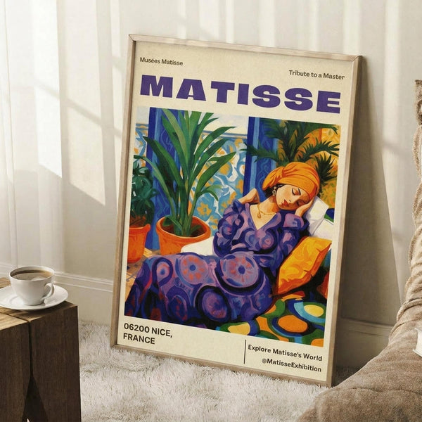 May include: Framed Matisse exhibition poster featuring a woman in purple patterned dress with orange headwrap, reclining among tropical plants in terracotta pots. Vibrant blues, greens, oranges in fauve style. Cream background with "MATISSE" in bold purple letters, "06200 NICE, FRANCE" at bottom. Light wood frame leaning against white wall next to beige sofa, with coffee cup on wooden side table and white textured rug.
