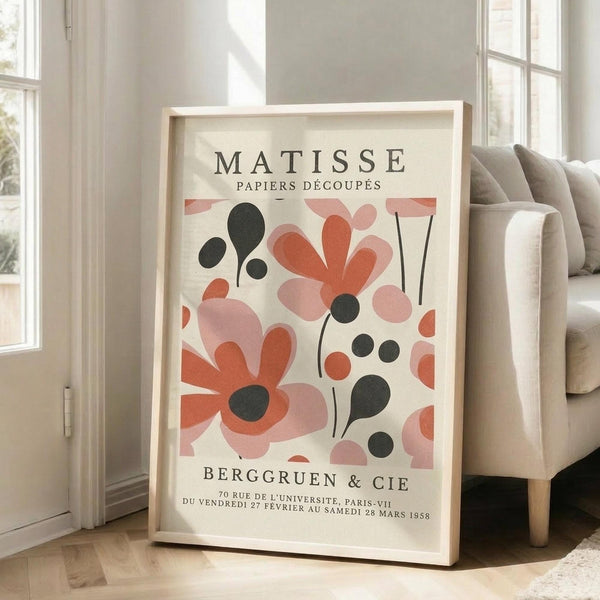 May include: Framed vintage exhibition poster with abstract floral cutout shapes in coral pink, salmon, and black on cream background, leaning against white wall next to beige upholstered sofa in bright living room with large windows and natural light streaming across hardwood floors.