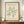 May include: Framed poster with sage green coral-like organic shapes on cream background, displayed on wooden sideboard. Brown wooden frame, accompanied by white ceramic vase and brass lantern on books. Neutral wall with white molding visible in background.