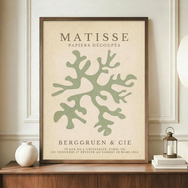 May include: Framed poster with sage green coral-like organic shapes on cream background, displayed on wooden sideboard. Brown wooden frame, accompanied by white ceramic vase and brass lantern on books. Neutral wall with white molding visible in background.