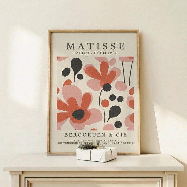 May include: Framed vintage-style art print featuring abstract cut-out shapes in coral pink, terracotta, and black against cream background, displayed on light wood console table with small wrapped gift box beside it on white wall