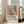 May include: Framed art poster displaying a colorful still life scene with geometric shapes, flowers in a vase, fruit bowl, and window view in muted oranges, blues, and greens. Light wooden frame leans against white wall beside beige sofa in bright living room with large windows and natural lighting.