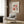 May include: Framed Matisse-inspired botanical poster with coral, pink and black cut-out style flowers hanging on wood paneled wall above beige upholstered armchair with geometric patterned pillow, brass floor lamp with white marble base, and dark round side table with open book in modern living room setting