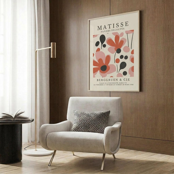 May include: Framed Matisse-inspired botanical poster with coral, pink and black cut-out style flowers hanging on wood paneled wall above beige upholstered armchair with geometric patterned pillow, brass floor lamp with white marble base, and dark round side table with open book in modern living room setting