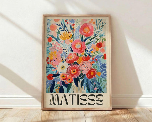 May include: A framed floral art print leaning against a white wall on light wooden flooring. The artwork features vibrant watercolor-style flowers in coral, pink, orange, and yellow with blue and green foliage against a cream background. The word "MATISSE" appears in bold black letters at the bottom. Natural light casts soft shadows across the scene, and the print is displayed in a simple light wood frame.