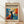 May include: A framed art poster leaning against a white wall on light wood flooring. The poster features a stylized black silhouette of a person with arms raised, surrounded by vibrant blue, orange, red and yellow abstract foliage shapes in a Matisse-inspired cut-out style. The design has a cream background with bold typography reading "matisse modernist" at the top and event details. Natural light creates soft shadows on the white fabric backdrop.