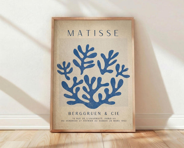 May include: Framed vintage exhibition poster with blue coral-like branching forms on cream background, displayed in light wood frame against white fabric backdrop on wooden floor. Poster features "MATISSE" text at top and "BERGGRUEN & CIE" with French gallery details below the organic blue shapes.
