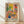 May include: Framed poster print in light wood frame leaning against white wall on wooden floor. Artwork shows colorful Matisse-style interior scene with black and white patterned chair, blue vase with plants, orange flowers, yellow door or window, pink tiled wall with small artworks, and tropical foliage. Features vibrant colors including yellows, pinks, greens, and blues in flat, painterly style with "MATISSE" text at bottom.