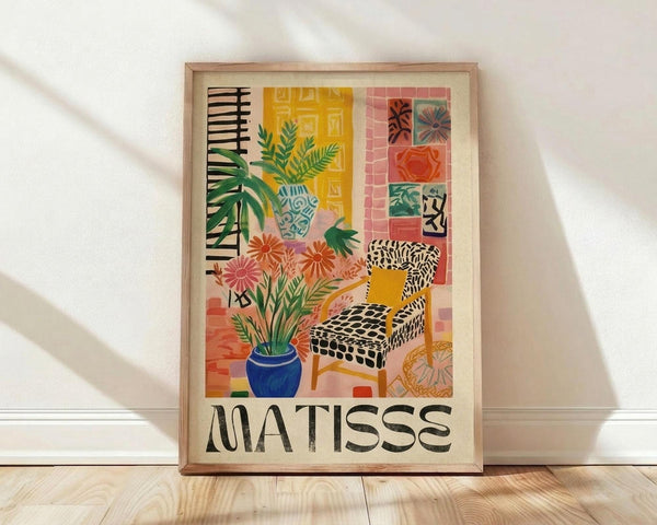 May include: Framed poster print in light wood frame leaning against white wall on wooden floor. Artwork shows colorful Matisse-style interior scene with black and white patterned chair, blue vase with plants, orange flowers, yellow door or window, pink tiled wall with small artworks, and tropical foliage. Features vibrant colors including yellows, pinks, greens, and blues in flat, painterly style with "MATISSE" text at bottom.