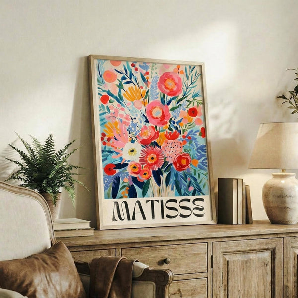 May include: Colorful floral art print with vibrant pink, orange, and blue flowers in Matisse's distinctive style, displayed in light wood frame on rustic wooden sideboard alongside fern plant, ceramic vase, and stacked books in cozy living room setting with cream upholstered furniture and warm natural lighting.