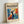 May include: Framed poster displaying modernist artwork with vibrant blue, orange, and red abstract shapes surrounding a black silhouette figure with raised arms. Text reads "matisse modernist" at top and "Embracing Colorful Freedom In the Spirit of Matisse" at bottom. Light wood frame sits against white wall on white surface.