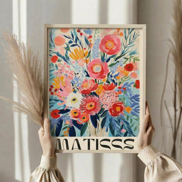 May include: Person holding framed floral art print in light wood frame against white curtained window. Artwork features vibrant bouquet with pink, coral, yellow and white flowers, blue and green foliage in painterly style with "MATISSE" text below. Dried wheat stems visible beside frame. Person wears cream ruffled sleeves.