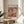 May include: Framed Matisse cut-out style art print displayed on round wooden side table next to beige upholstered chair. Artwork features abstract organic shapes in warm oranges, deep reds, yellows and cream with crosshatched patterns. Natural lighting creates soft shadows across neutral room setting with plant visible in corner.