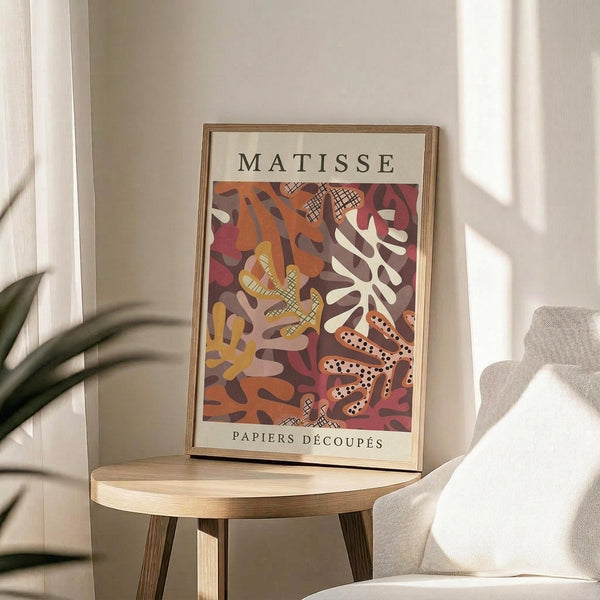 May include: Framed Matisse cut-out style art print displayed on round wooden side table next to beige upholstered chair. Artwork features abstract organic shapes in warm oranges, deep reds, yellows and cream with crosshatched patterns. Natural lighting creates soft shadows across neutral room setting with plant visible in corner.