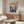 May include: Colorful modernist poster featuring a silhouetted figure with raised arms surrounded by vibrant blue, orange, and red abstract shapes, displayed in a thin wooden frame above a walnut credenza. Scene shows minimalist living room with white curved chair, glass oval coffee table, and warm natural lighting creating soft shadows on cream walls.