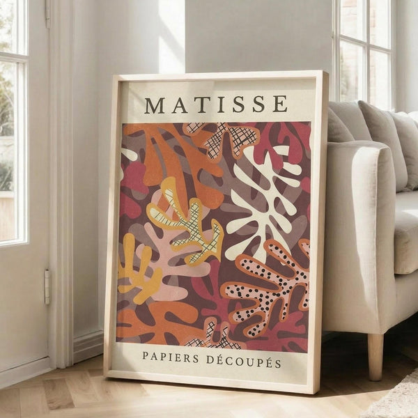 May include: A framed art print leaning against a white wall beside a beige upholstered sofa. The poster features abstract cut-out shapes in warm tones of orange, burgundy, yellow, and cream with cross-hatched patterns and organic coral-like forms. White text reads "MATISSE" at top and "PAPIERS DÉCOUPÉS" at bottom. Natural sunlight streams through tall windows onto hardwood floors in this minimalist living room setting.