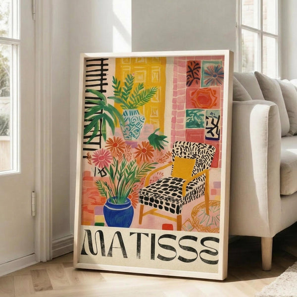 May include: Colorful Matisse-inspired poster in white frame leaning against wall next to beige sofa. Artwork features vibrant interior scene with patterned chair, blue vase with palm fronds, tropical plants, and geometric wall tiles in yellow, pink, and coral tones. Bold black "MATISSE" text at bottom.