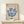May include: Framed vintage-style exhibition poster featuring blue coral-like branching forms on cream background, displayed on light wood console table beside small white gift box with green twine, against neutral wall with natural lighting