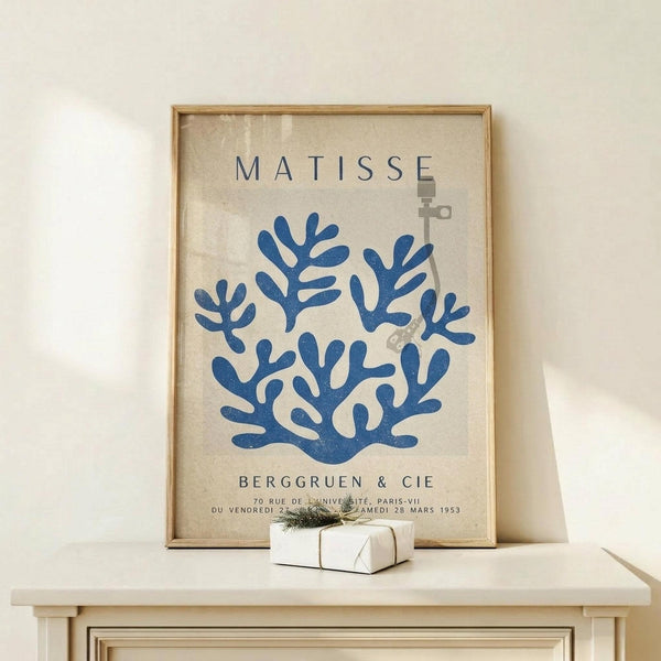 May include: Framed vintage-style exhibition poster featuring blue coral-like branching forms on cream background, displayed on light wood console table beside small white gift box with green twine, against neutral wall with natural lighting