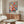 May include: Colorful floral art print with vibrant pink, orange, and blue flowers in an abstract style, displayed in a thin frame above a walnut wood credenza. Modern living room setting features a white curved chair, glass-top oval coffee table, and warm neutral walls with natural lighting.