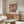 May include: Framed vintage-style art poster featuring Matisse's colorful cut-paper collage design with abstract leaf and coral shapes in warm oranges, reds, yellows and browns, displayed above a mid-century modern walnut credenza in a minimalist living room with white curved chair, glass coffee table, and brass floor lamp.