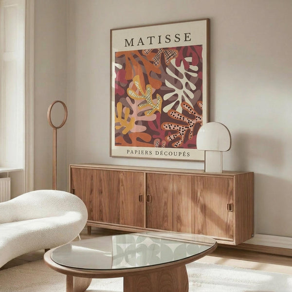May include: Framed vintage-style art poster featuring Matisse's colorful cut-paper collage design with abstract leaf and coral shapes in warm oranges, reds, yellows and browns, displayed above a mid-century modern walnut credenza in a minimalist living room with white curved chair, glass coffee table, and brass floor lamp.