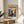 May include: Framed poster with modernist design featuring a silhouetted figure with raised arms surrounded by vibrant blue and orange abstract shapes, displayed on a light wood round side table next to a beige upholstered chair. Natural sunlight streams across white walls, with a plant's shadow visible in the corner of this minimalist living room setting.