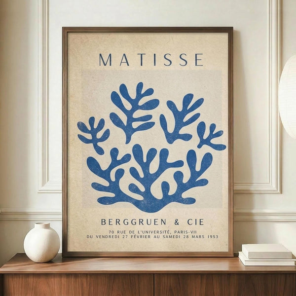 May include: Framed vintage exhibition poster featuring blue coral-like seaweed forms on cream textured background, displayed on wooden sideboard with small white ceramic vase and stacked books in elegant room with white paneled walls.