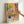 A colorful art print in a light wood frame held by a hand against a white wall. The artwork depicts a vibrant interior scene with a patterned armchair, potted plants including flowers in a blue vase and leafy plants in decorative vessels, geometric wall tiles in coral and yellow tones, and abstract patterns in black, white, and bright colors. The word "MATISSE" appears at the bottom in bold black letters. The style features bold colors, simplified forms, and decorative patterns reminiscent of mi