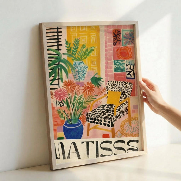 A colorful art print in a light wood frame held by a hand against a white wall. The artwork depicts a vibrant interior scene with a patterned armchair, potted plants including flowers in a blue vase and leafy plants in decorative vessels, geometric wall tiles in coral and yellow tones, and abstract patterns in black, white, and bright colors. The word "MATISSE" appears at the bottom in bold black letters. The style features bold colors, simplified forms, and decorative patterns reminiscent of mi