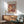 May include: Framed Matisse-style poster with colorful abstract cut-out shapes in warm oranges, reds, and earth tones, displayed above a mid-century modern wooden credenza with white ceramic lamp. Sage green upholstered armchair with wooden frame sits nearby in sunlit room with white walls and wooden floors.