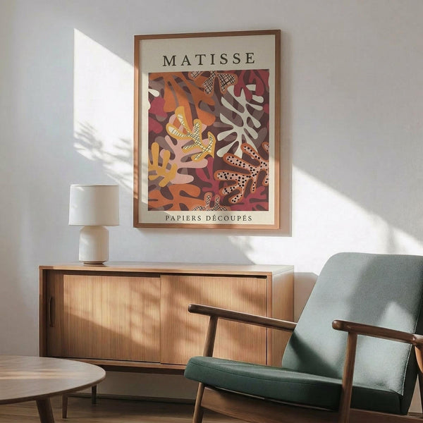 May include: Framed Matisse-style poster with colorful abstract cut-out shapes in warm oranges, reds, and earth tones, displayed above a mid-century modern wooden credenza with white ceramic lamp. Sage green upholstered armchair with wooden frame sits nearby in sunlit room with white walls and wooden floors.
