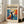 May include: Framed poster displaying abstract modernist artwork with vibrant blue, orange, and yellow colors featuring a black silhouette figure with raised arms surrounded by leaf-like shapes. The cream-colored poster shows exhibition text at top and bottom. White wooden frame leans against beige sofa in bright sunlit room with white walls and hardwood floors.