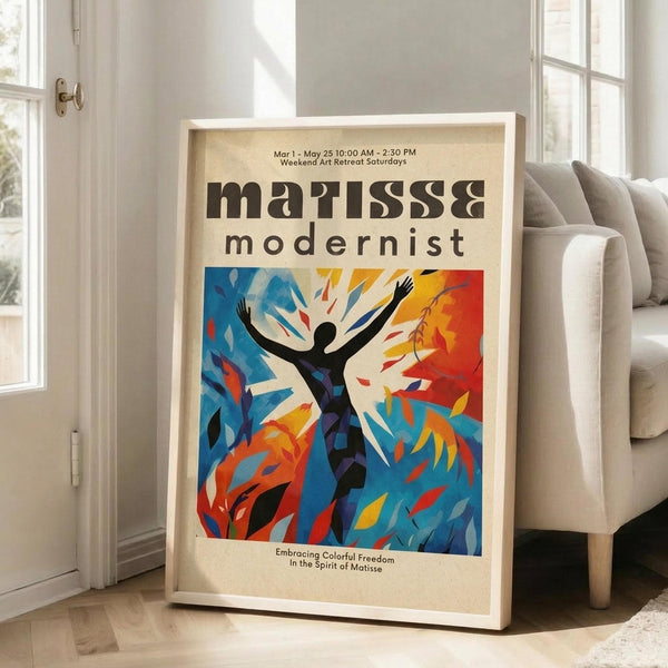 May include: Framed poster displaying abstract modernist artwork with vibrant blue, orange, and yellow colors featuring a black silhouette figure with raised arms surrounded by leaf-like shapes. The cream-colored poster shows exhibition text at top and bottom. White wooden frame leans against beige sofa in bright sunlit room with white walls and hardwood floors.