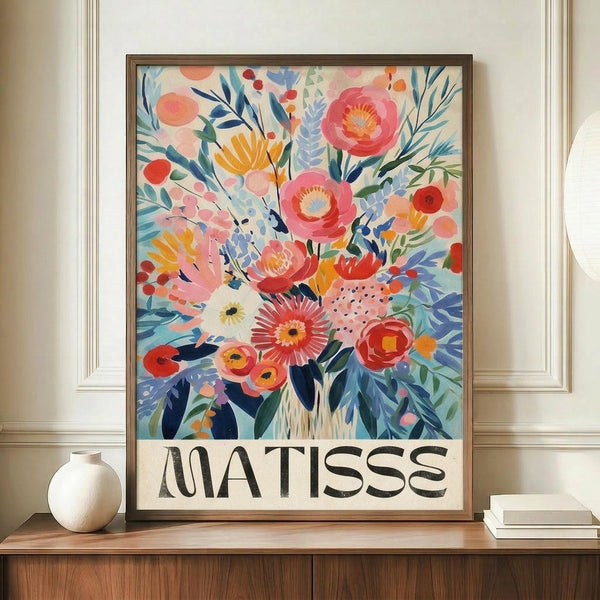 May include: Vibrant floral poster in warm wooden frame displaying colorful abstract flowers in pink, coral, yellow, and blue with botanical leaves and stems against cream background, with "MATISSE" text at bottom. Artwork sits on light wood sideboard next to white ceramic vase and stacked books in modern interior with white paneled walls.