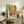 May include: Framed Matisse-style art print displayed on rustic wooden sideboard in warm-toned living room. Colorful poster features interior scene with tropical plants in blue vase, patterned armchair, geometric tiles in coral and yellow tones. Natural wood frame complements surrounding decor including fern plant, ceramic vase, and cream upholstered furniture. Soft natural lighting creates cozy atmosphere.