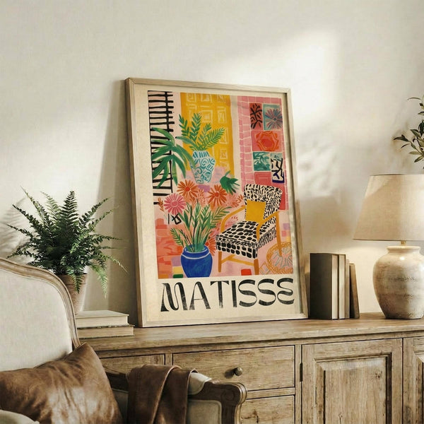 May include: Framed Matisse-style art print displayed on rustic wooden sideboard in warm-toned living room. Colorful poster features interior scene with tropical plants in blue vase, patterned armchair, geometric tiles in coral and yellow tones. Natural wood frame complements surrounding decor including fern plant, ceramic vase, and cream upholstered furniture. Soft natural lighting creates cozy atmosphere.
